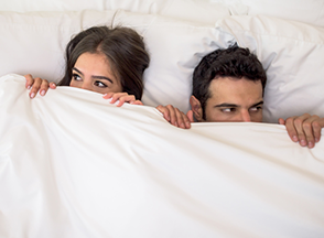 A man and woman in bed with white bed sheet up to their nose.